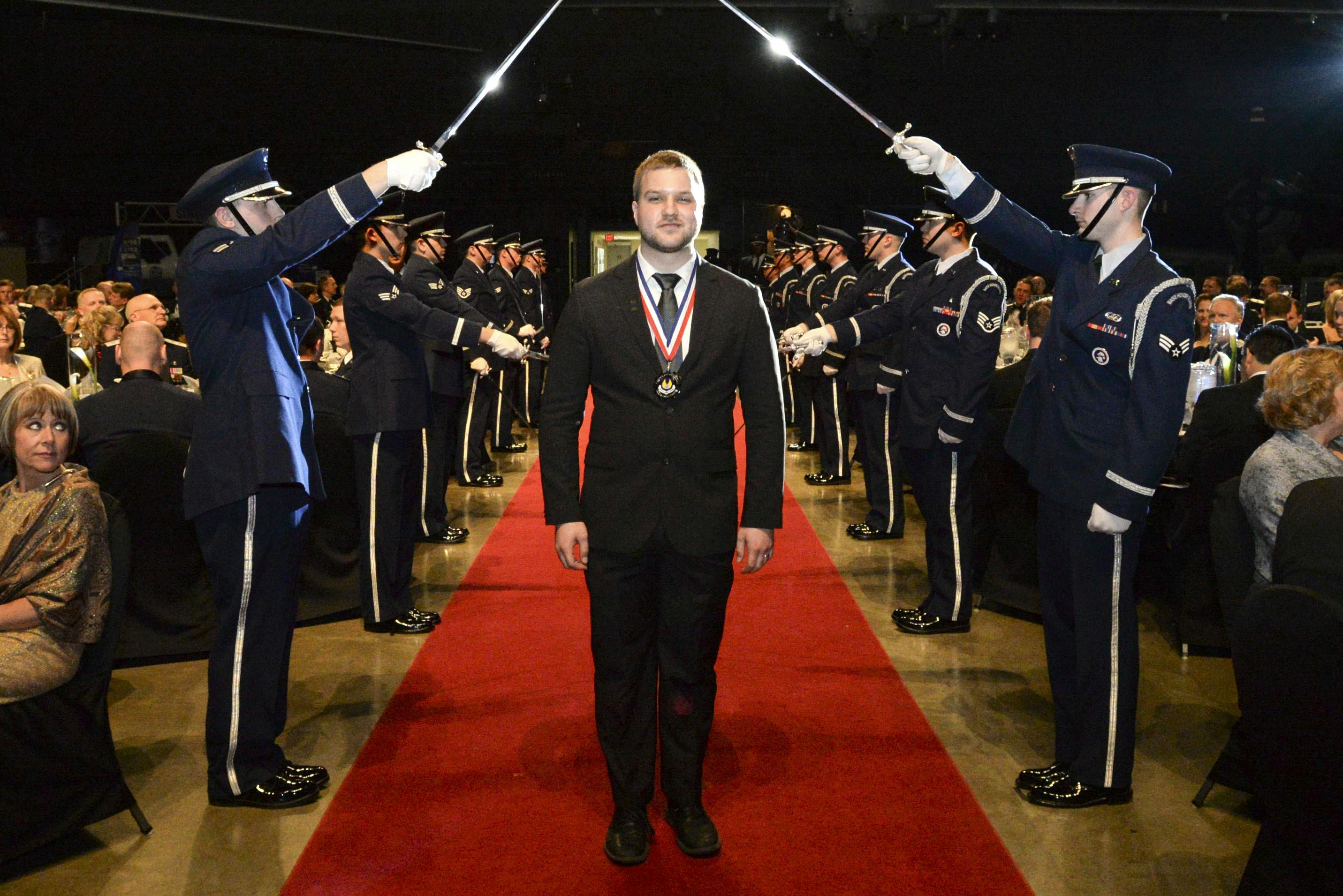 Ty Mick, wearing a medal featuring the AFMC logo, walking on a red carpet between Airmen holding swords at the 2014 AFMC Annual Excellents Awards.